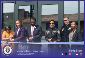 A group of individuals stand in front of a building entrance during a ribbon‑cutting ceremony. Several people hold large scissors and cut a wide blue ribbon stretched across the scene. The individuals are dressed in formal clothing, including business attire and police uniforms with badges and patches. Behind them is a modern glass‑door entrance and part of a building with a brown roof. At the bottom of the image, a Memphis Police Department emblem appears on the left with the text “Memphis Police Department – Established in 1827.” On the right side are icons for social media and the website address “joinmpd.com,” all within a blue border.