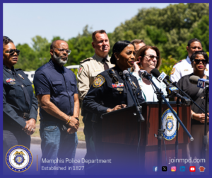 A group of uniformed and non‑uniformed individuals stand outdoors behind a podium during a press event. Several microphones from different news outlets are positioned on the podium, which displays the Memphis Police Department seal. The group stands in a row with trees and greenery visible in the background. At the bottom of the image, text reads “Memphis Police Department – Established in 1827” along with social media icons and the website joinmpd.com.