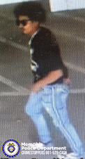 A surveillance-style image shows an individual moving through a parking area with visible painted lines on the ground. The individual is wearing a dark short-sleeved top with a graphic on the front, light-colored jeans with vertical white stripes along the sides, and athletic shoes with contrasting colors. The background appears to be part of a covered or indoor parking structure. In the lower left corner, a Memphis Police Department badge graphic appears with text that reads “Memphis Police Department CRIMESTOPPERS 901‑528‑CASH.”