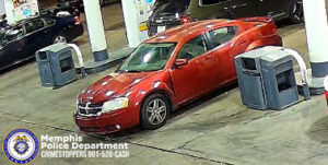 The image shows a red four‑door sedan parked beside a gas pump at a fuel station. The car is positioned at an angle with the front end pointed slightly toward the left side of the image. Multiple gas pumps with white and blue color accents are visible around the vehicle. Additional parked vehicles can be seen in the background, including a dark‑colored sedan on the left. The ground surface is concrete with visible wear marks. At the bottom left corner of the image is the Memphis Police Department seal and text that reads “Memphis Police Department CRIMESTOPPERS 901‑528‑CASH.”
