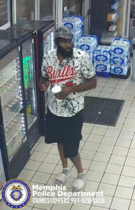 The image shows an individual walking inside what appears to be a convenience store. The person is wearing a short‑sleeve shirt with a white, black, and red graphic design featuring the word “Bulls” in large red letters across the chest, along with dark knee‑length shorts and light‑colored shoes. The individual is holding a white cup and a small object in the other hand. To the left are glass‑front refrigerator units with beverages inside. In the background are stacks of packaged bottled water arranged against the wall. The floor consists of light‑colored tiles with dark mats placed near the entrance. At the bottom of the image is the Memphis Police Department seal along with white text that reads “Memphis Police Department CRIMESTOPPERS 901‑528‑CASH.”