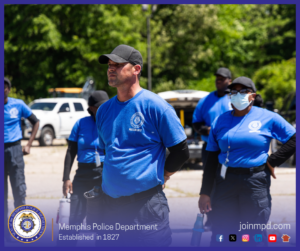 Several individuals wearing blue shirts labeled “Recruit” stand outdoors in a training or instructional setting. The group appears to be in a parking or staging area with vehicles and trees in the background. Some individuals hold water bottles, and the scene is brightly lit by sunlight. At the bottom of the image, text displays “Memphis Police Department – Established in 1827,” along with various social media icons and the website joinmpd.com.