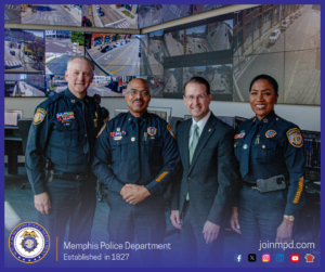 Four individuals stand together inside a control‑room environment filled with multiple computer monitors and large wall‑mounted screens showing various live or recorded street and traffic camera views. Three individuals are wearing Memphis Police Department uniforms with visible patches, badges, nameplates, and service decorations. One individual is dressed in a business suit. The group stands closely in front of several workstations where additional people are seated at computers. The bottom portion of the image includes a Memphis Police Department emblem on the left, the text “Memphis Police Department – Established in 1827,” and the website “joinmpd.com” with several social media icons on the right, all framed by a blue border.