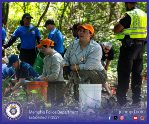 A group of individuals are gathered in a wooded outdoor area, participating in what appears to be a hands‑on training or search activity. Several people wearing blue shirts are positioned throughout the background, while two individuals in light‑colored shirts and orange caps are kneeling or sitting on large plastic buckets in the foreground. Buckets, tools, and containers are scattered around the area. On the right side of the image, a uniformed person wearing a high‑visibility vest stands observing the scene. Sunlight filters through the trees, illuminating patches of foliage and casting shadows on the ground. At the bottom of the image, text reads “Memphis Police Department – Established in 1827,” along with social media icons and the website joinmpd.com.
