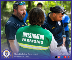 A group of individuals are gathered outdoors in a wooded area during what appears to be an investigative or training activity. At the center of the image, a person wearing a green vest with bold white text reading “INVESTIGATOR FORENSICS” stands with their back to the camera. Surrounding this individual are others wearing dark shirts and uniforms, some featuring patches or insignias on the sleeves or chest. Sunlight filters through the trees, casting a mix of light and shadow across the scene. At the bottom of the image, text reads “Memphis Police Department – Established in 1827,” along with social media icons and the website joinmpd.com.
