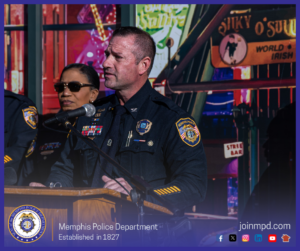 Colonel Penny stands at a podium equipped with a microphone, delivering remarks during an event. Other uniformed personnel stand nearby. The background features a colorful, illuminated streetscape-style backdrop with various signs, including one that reads “Silky O’Sullivan’s World Famous Irish Bar” and another reading “Strip Bar.” The individuals are wearing formal police uniforms with department patches, badges, and service decorations. At the bottom of the image, a Memphis Police Department emblem is displayed alongside the text “Memphis Police Department – Established in 1827,” with the website “joinmpd.com” and several social media icons on the right, all within a blue border.