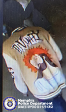 A surveillance-style image shows an individual from the back wearing a light-colored varsity-style jacket. The jacket has large lettering across the upper back and a large graphic below it depicting a stylized figure with bright orange hair and hands raised near the face. The individual is standing indoors on a tiled floor, and parts of other individuals are partially visible around the frame. In the lower left corner, a Memphis Police Department badge graphic appears along with text that reads “Memphis Police Department CRIMESTOPPERS 901‑528‑CASH.”