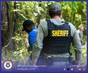  A person wearing tactical gear with a vest labeled “SHERIFF” in large yellow letters stands in a wooded area. The individual is facing away from the camera and appears to be observing or accompanying another person in a blue shirt who is slightly ahead and to the left in the trees. Dense foliage, tree trunks, and patches of sunlight create a forested backdrop. At the bottom of the image, text reads “Memphis Police Department – Established in 1827,” along with social media icons and the website joinmpd.com.