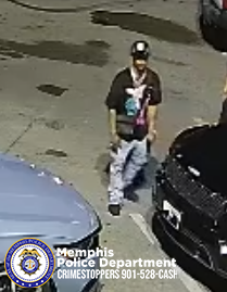 A surveillance-style image shows one person standing outdoors in a parking lot at night. The person is positioned between two parked vehicles, one light‑colored and one dark‑colored. The person is wearing a dark shirt with a large graphic on the front, light-colored pants, a hat, and a crossbody-style bag. The ground is wet and several dark patches are visible on the pavement. In the bottom left corner, a Memphis Police Department badge graphic is displayed along with text that reads “Memphis Police Department CRIMESTOPPERS 901‑528‑CASH.”