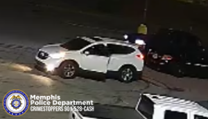 A surveillance-style image shows a white SUV parked in a lot at night with the front driver-side door open. A dark-colored vehicle is parked nearby, and a person wearing light and dark clothing stands beside it. In the background, a person wearing a bright, light-colored top is visible near the edge of the lot. The pavement appears worn and cracked. In the bottom left corner, a Memphis Police Department badge graphic is displayed along with text that reads “Memphis Police Department CRIMESTOPPERS 901‑528‑CASH.”