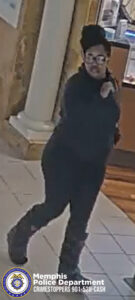 The image shows an individual walking inside a building with tiled flooring, possibly a shopping mall or similar indoor public space. The person is wearing dark clothing, including a long‑sleeve top, pants, and footwear with a textured or rugged design. A dark hat or head covering is also worn. The individual appears to be in mid‑step and is holding an object in one hand. In the background, there is a large decorative column, part of a wall with ornate detailing, and what appears to be a counter or display area with various items visible. At the bottom left corner of the image is the Memphis Police Department seal and white text reading “Memphis Police Department CRIMESTOPPERS 901‑528‑CASH.”