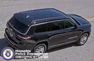 The image shows a dark-colored Jeep Cherokee SUV captured from an elevated angle in daylight. The vehicle is positioned on a paved roadway with a faint yellow centerline visible beneath it. The SUV appears to be in motion or recently moving, and its windows, roof rails, and body contours are clearly visible. Shadows cast by the vehicle fall toward the lower-right side of the image. In the lower-left corner, the Memphis Police Department seal is present along with white text that reads “Memphis Police Department CRIMESTOPPERS 901‑528‑CASH.”