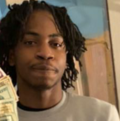  A person, the homicide victim, with medium-length, twisted hair is shown indoors, wearing a light-colored shirt. The individual is holding a visible stack of paper currency in one hand. A wooden doorframe and a partially painted wall are in the background.
