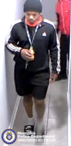 A person wearing a black athletic jacket with white stripes on the sleeves, black shorts, white socks, and dark slide-style footwear is walking down a narrow hallway. The individual holds a drink bottle and a lanyard. Another person in a red jacket is visible standing further down the hallway. At the bottom of the image, the Memphis Police Department badge and the text “Memphis Police Department CRIMESTOPPERS 901-528-CASH” are displayed.