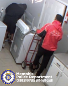 Two individuals are inside a small laundry or utility room with white cabinets and light-colored tile flooring. One individual wearing dark clothing is positioned near a washing machine, leaning over it. The other individual, wearing a red jacket with black sleeves and dark pants, is holding a red hand truck that is positioned next to the washing machine. The scene is captured from an overhead angle. At the bottom of the image, the Memphis Police Department badge and the text “Memphis Police Department CRIMESTOPPERS 901‑528‑CASH” are visible.