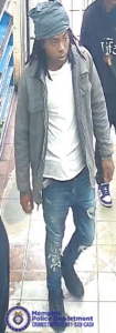 A surveillance-style image taken inside a store shows one person walking on a tiled floor. The person is wearing a head covering, a light-colored shirt, a zip-up jacket, distressed jeans, and dark shoes. Another person is partially visible behind them, wearing dark clothing and sneakers. Shelving stocked with items lines the left side of the image. In the bottom left corner, a Memphis Police Department badge graphic appears alongside text that reads “Memphis Police Department CRIMESTOPPERS 901‑528‑CASH.”