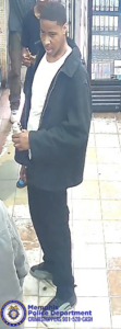 A surveillance-style image taken inside a store shows one person standing on a tiled floor while holding an item in one hand. The person is wearing a dark jacket, a light-colored shirt, dark pants, and light-colored shoes. Behind this individual, another person is partially visible wearing dark clothing and sneakers. Refrigerated display cases with rows of beverages line the right side of the image, while shelving is visible on the left. In the bottom left corner, a Memphis Police Department badge graphic appears along with text reading “Memphis Police Department CRIMESTOPPERS 901‑528‑CASH.”