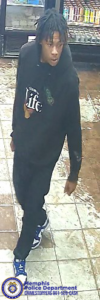 A surveillance-style image taken inside a store shows one individual walking across a tiled floor. The individual is wearing a dark hooded sweatshirt with a graphic on the front, dark pants, and athletic shoes with light and dark contrasting colors. Refrigerated shelves filled with assorted products line the background. The floor has a mix of light and darker tiles, some appearing worn. In the bottom left corner, a Memphis Police Department badge graphic appears with text that reads “Memphis Police Department CRIMESTOPPERS 901‑528‑CASH.”