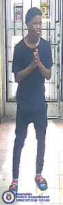 A surveillance-style image taken inside a store shows one individual standing on a tiled floor with hands held together in front. The individual is wearing a dark short-sleeved shirt, dark pants, and slide-style sandals with brightly colored straps. The background includes a metal security gate and glass door with various posted signs. To the left edge of the image, part of another person is visible. In the bottom left corner, a Memphis Police Department badge graphic appears alongside text that reads “Memphis Police Department CRIMESTOPPERS 901‑528‑CASH.”