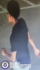 A surveillance-style image shows an individual moving through a parking area marked with painted lines. The individual is wearing a dark short-sleeved shirt and light-colored pants. The ground surface appears textured, and the lighting suggests the area may be partially covered. In the lower left corner, a Memphis Police Department badge graphic appears with text that reads “Memphis Police Department CRIMESTOPPERS 901‑528‑CASH.”