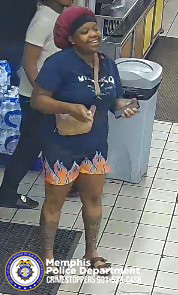 The image shows an individual standing inside what appears to be a convenience store. The person is wearing a dark short‑sleeve shirt with white text on the front, patterned shorts with a flame‑style design near the hem, and light‑colored footwear. The individual is holding objects in both hands, and a bag or pouch is visible at the waist. In the background, there are stacks of packaged bottled water, a trash bin, tiled flooring, and part of a store counter. At the bottom of the image is the Memphis Police Department seal along with white text that reads “Memphis Police Department CRIMESTOPPERS 901‑528‑CASH.”