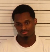 A person is standing in front of a height‑measurement backdrop consisting of horizontal black lines on a light background. The individual is wearing a plain light-colored shirt, and the shoulders and upper torso are visible.