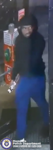 A surveillance-style image shows a person wearing a dark hooded jacket and bright blue pants standing near the entrance of a store or gas station. The person is holding an object in one hand that appears to be rectangular and mostly white. Behind the individual is a fuel pump with red and green circular graphics. In the lower-left corner, the Memphis Police Department seal and the text “Memphis Police Department CRIMESTOPPERS 901-528-CASH” are visible.