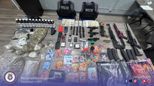 "A large evidence display is arranged on a table in a police office setting. The items include multiple firearms, rifle magazines, and ammunition; several bags filled with what appears to be plant material packaged in different quantities; numerous bottles labeled with numbers; various small electronic devices including mobile phones and digital scales; stacks of cash; several bags containing colorful packaged products resembling edibles or candy‑style items; two tactical vests labeled “Memphis Police Fugitive Unit”; and assorted containers and paraphernalia. The table is positioned against a wall with electrical outlets and office furniture in the background. At the bottom of the image is a Memphis Police Department banner with the department seal, the text “Memphis Police Department Established in 1827,” and the website “joinmpd.com,” along with social media icons."