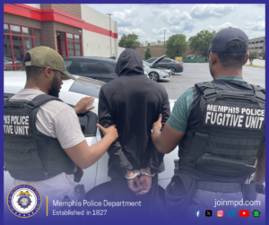 Two individuals wearing tactical vests labeled “MEMPHIS POLICE FUGITIVE UNIT” are standing on either side of another individual who is wearing a hooded sweatshirt and has hands secured behind the back with handcuffs. All three are positioned near the rear of a white vehicle in a parking lot. Several parked vehicles, a building with a red and white exterior, trees, and a partly cloudy sky appear in the background. At the bottom of the image, there is a Memphis Police Department seal along with the text “Memphis Police Department — Established in 1827” and “joinmpd.com,” followed by various social media icons.