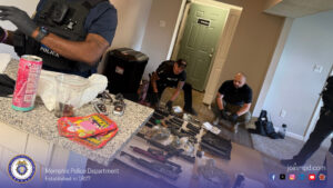 A law‑enforcement evidence scene inside a residential interior. Several officers wearing tactical gear are present, with one standing in the left foreground near a counter and two others kneeling on the floor in the background. Numerous items are laid out on the floor, including multiple firearms, firearm magazines, ammunition, clear plastic bags containing plant material, and other evidence. The countertop in the foreground holds a tall beverage can, a clear plastic cup, and brightly colored packaged items. A green door with a sign is visible in the background, along with a large black bin against the wall. At the bottom of the image, a Memphis Police Department banner displays the department seal, the text “Memphis Police Department Established in 1827,” the website “joinmpd.com,” and several social media icons.