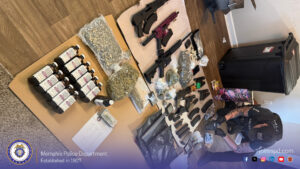 "A room scene showing a large evidence display arranged on the floor and on cardboard panels. The items include multiple firearms in different sizes and colors, several magazines and ammunition, sealed bags containing plant material, numerous bottles with printed labels, small electronic devices, a stack of documents, and various other packaged items. A person wearing tactical gear is positioned on the right side of the image, kneeling and writing on paperwork. The floor is wood‑patterned, and a large black bin is placed against the wall. At the bottom of the image, a Memphis Police Department banner is visible with the department seal, the text “Memphis Police Department Established in 1827,” and the website “joinmpd.com,” along with social media icons."