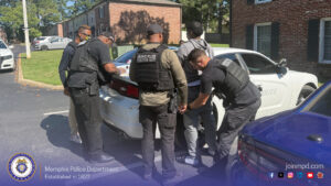 Several law‑enforcement officers wearing tactical gear are gathered around a white police vehicle in an outdoor residential parking area. One individual is positioned against the rear of the vehicle while officers stand close, appearing to conduct an inspection or procedure. The scene takes place in front of brick apartment buildings with green lawns and trees in the background. Additional police vehicles are parked nearby, including a blue one partly visible on the right. At the bottom of the image, a Memphis Police Department banner displays the department seal, the text “Memphis Police Department Established in 1827,” the website “joinmpd.com,” and social media icons.