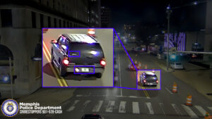 A nighttime street scene from a surveillance camera showing a vehicle driving down a multi‑lane road lined with construction barrels. Buildings and streetlights are visible in the background. A dark-colored SUV is traveling near the center of the road, and a zoomed‑in inset highlights the rear of the same vehicle. The inset includes purple boxes around two areas: one on the back window and one around the license plate area. The Memphis Police Department seal and the text “Memphis Police Department CRIMESTOPPERS 901‑528‑CASH” appear in the lower-left corner of the image.