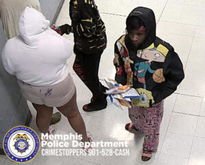 Three individuals are standing in an indoor hallway with light-colored tile flooring and light walls. One individual in the foreground is wearing a hooded jacket featuring a colorful cartoon-style design, patterned pants, and open-toed footwear while holding several small packaged items. Another individual nearby is wearing a light-colored hooded top and shorts with visible markings on the back. A third individual is wearing a dark hooded top with a pattern and dark shorts. In the lower-left corner, a Memphis Police Department badge and the text “Memphis Police Department CRIMESTOPPERS 901‑528‑CASH” are displayed.