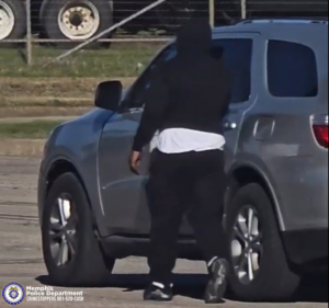 An individual wearing a dark hooded top, dark pants, and light‑soled footwear is walking toward the driver side of a gray SUV parked outdoors. A white shirt is visible beneath the dark outer clothing. The vehicle is positioned on a paved surface near a fenced area with large vehicle tires stacked in the background. In the lower‑left corner, a Memphis Police Department badge logo appears next to text reading “Memphis Police Department CRIMESTOPPERS 901‑528‑CASH.”