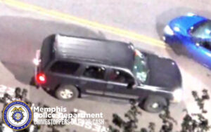 An overhead nighttime image showing a dark-colored SUV driving on a roadway. The SUV is positioned near the center of the image, traveling alongside a yellow centerline. A blue car is partially visible at the top right. In the bottom left corner, the Memphis Police Department seal and the text “Memphis Police Department CRIMESTOPPERS 901‑528‑CASH” are displayed. Some tree branches appear in the lower foreground, partially obscuring the view.