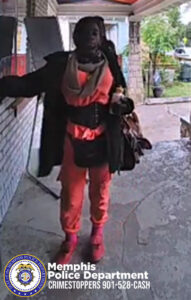 An individual stands on a covered porch next to a brick wall, wearing a long dark coat layered over a pink outfit. The individual has a large scarf wrapped around the neck area, a wide belt around the waist, and carries a large handbag on the right arm. The person is positioned near a white door and appears to be extending one arm toward the wall. The porch has a textured concrete floor, and in the background there is a yard with plants and a small animal visible near the edge of the frame. In the lower-left corner, there is a Memphis Police Department CrimeStoppers logo and the text 'Memphis Police Department CRIMESTOPPERS 901-528-CASH.'