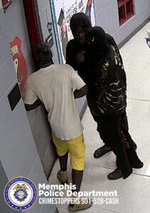 Three individuals are standing close together in a hallway with light-colored tile flooring and light walls. One individual, wearing a light-colored T‑shirt and bright yellow shorts, is facing a closed door and appears to be interacting with something near the door’s handle area. Another individual, wearing a dark hooded top with a pattern and dark pants, is positioned slightly behind and to the side. A third individual, wearing a dark hooded top and dark pants, is standing close to the group. The hallway contains posters and artwork on the walls. In the lower-left corner, a Memphis Police Department badge and text reading “Memphis Police Department CRIMESTOPPERS 901‑528‑CASH” are visible.