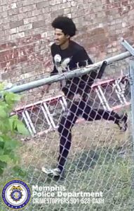 A person is captured mid‑stride while running alongside a chain‑link fence in an outdoor area with grass and scattered vegetation. The person is wearing dark clothing, including a long‑sleeved shirt with a large graphic on the front and dark pants. A ladder is visible on the ground behind the fence, leaning against a brick wall in the background. In the lower-left corner, there is a Memphis Police Department and CrimeStoppers information badge and text.