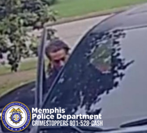 An individual is standing beside a dark-colored vehicle near the front passenger-side door. The person is partially visible, with the vehicle occupying much of the right side of the image. Trees, grass, and a paved surface are visible in the background. In the lower‑left corner, there is a Memphis Police Department badge logo along with the text “Memphis Police Department CRIMESTOPPERS 901‑528‑CASH.”