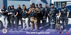 A group of individuals wearing tactical gear and vests labeled “MEMPHIS POLICE” are standing together outdoors in front of a large industrial-style building with metal siding and rolling bay doors. Several individuals are holding equipment. One individual is positioned next to a working dog wearing a harness. Multiple police vehicles are parked behind the group. At the bottom of the image, a Memphis Police Department seal appears on the left, along with the text “Memphis Police Department — Established in 1827.” On the right side is the text “The MPD Fugitive Unit” and “joinmpd.com,” accompanied by various social media icons.