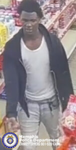 A person is walking down an aisle inside a store, surrounded by shelves stocked with various packaged goods. The person is wearing a light-colored sleeveless shirt, light-colored pants, and an open dark jacket. The individual is holding an item in one hand. In the lower-left corner of the image, a Memphis Police Department badge and text reading “Memphis Police Department CRIMESTOPPERS 901‑528‑CASH” are visible.