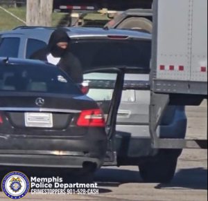 A dark-colored sedan with its rear door open is parked beside a larger vehicle, possibly a box truck. An individual wearing dark clothing and a hooded covering is standing near the open door of a silver SUV positioned in front of the sedan. The scene takes place outdoors on a paved surface in daylight. In the lower‑left corner, a Memphis Police Department badge logo appears with text reading “Memphis Police Department CRIMESTOPPERS 901‑528‑CASH.”