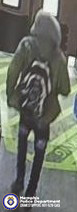 A surveillance‑style image taken inside a store. A person wearing a hooded jacket with the hood up, dark pants with worn or faded areas, and dark footwear is standing on a patterned floor near what appears to be a counter or store entrance area. The person is carrying a backpack with a bold circular or spiral design. In the bottom-left corner, a Memphis Police Department emblem is visible along with text reading “Memphis Police Department CRIMESTOPPERS 901‑528‑CASH.” Shelving, signage, and store fixtures are in the background.