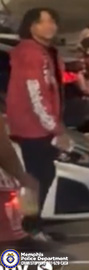 A person wearing a red long‑sleeved top with light-colored lettering on the sleeve is standing beside an open vehicle door in a dimly lit environment. The person is holding an object in one hand. Another individual is partially visible nearby. Red and white lights from the vehicle illuminate parts of the scene. A Memphis Police Department badge and CrimeStoppers contact information appear in the lower-left corner of the image.