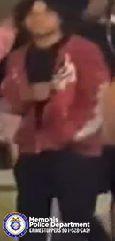 A person wearing a red long‑sleeved top with a white graphic design on the sleeves and chest is standing in a dimly lit setting among several other individuals. The person is holding a dark rectangular object with both hands in front of their torso. The background is somewhat blurred but shows additional people and soft lighting. In the lower-left corner, a Memphis Police Department badge and text reading “Memphis Police Department CRIMESTOPPERS 901‑528‑CASH” are visible.