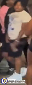 A person is standing among a group of other individuals in what appears to be a dimly lit or nighttime setting. The person is wearing a light-colored T‑shirt with a graphic design on the front, dark shorts, white shoes, and is holding an object near the waistband. Surrounding individuals are partially visible, with one wearing dark clothing and another wearing a bright pink top. In the lower-left corner, there is a Memphis Police Department badge and text that reads “Memphis Police Department CRIMESTOPPERS 901‑528‑CASH.”