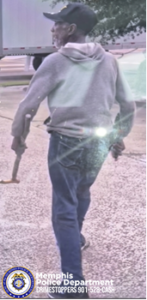 An individual wearing a light-colored hooded sweatshirt, dark pants, and a cap is shown walking outdoors on a paved surface. The individual is holding a long object in one hand. A large truck with a white trailer is visible ahead on the left side of the image, and trees and grass are in the background. A bright glare from sunlight appears near the center of the image. In the lower‑left corner, a Memphis Police Department badge logo appears with text reading “Memphis Police Department CRIMESTOPPERS 901‑528‑CASH.”