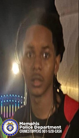 A person is shown in a dimly lit outdoor or nighttime setting, standing near a wall. The person is wearing a dark top with a red section visible on one side and a necklace around the neck area. Behind the person, there are bright lights, including a vertical arrangement of blue lights and a strong yellow light source. In the lower-left corner, the Memphis Police Department badge and text reading “Memphis Police Department CRIMESTOPPERS 901‑528‑CASH” are visible.