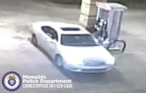 A grainy surveillance-style image shows a light-colored sedan parked next to a gas pump at night. The vehicle is positioned at an angle with its front facing slightly toward the camera. A person is standing beside the pump operating the fuel nozzle. The ground and surroundings are dimly lit. In the lower-left corner, there is a Memphis Police Department badge logo with white text reading “Memphis Police Department CRIMESTOPPERS 901-528-CASH.”